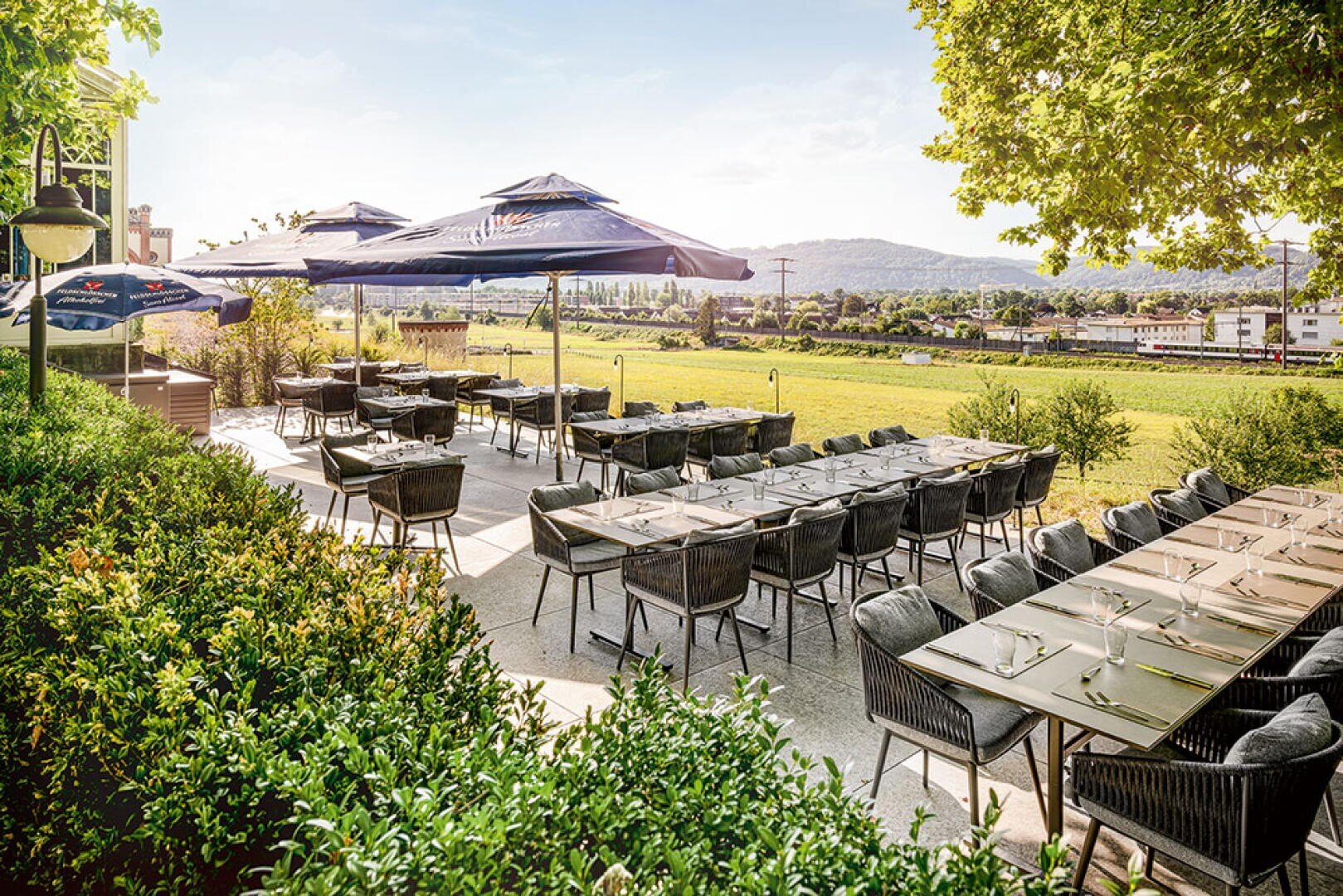 Restaurantterrasse im Freien mit Tischen und Stühlen zum Essen, beschattet von großen Sonnenschirmen. Grüne Büsche rahmen die Terrasse ein, mit einer Wiese, einem Fluss und Hügeln im Hintergrund unter einem sonnigen Himmel.