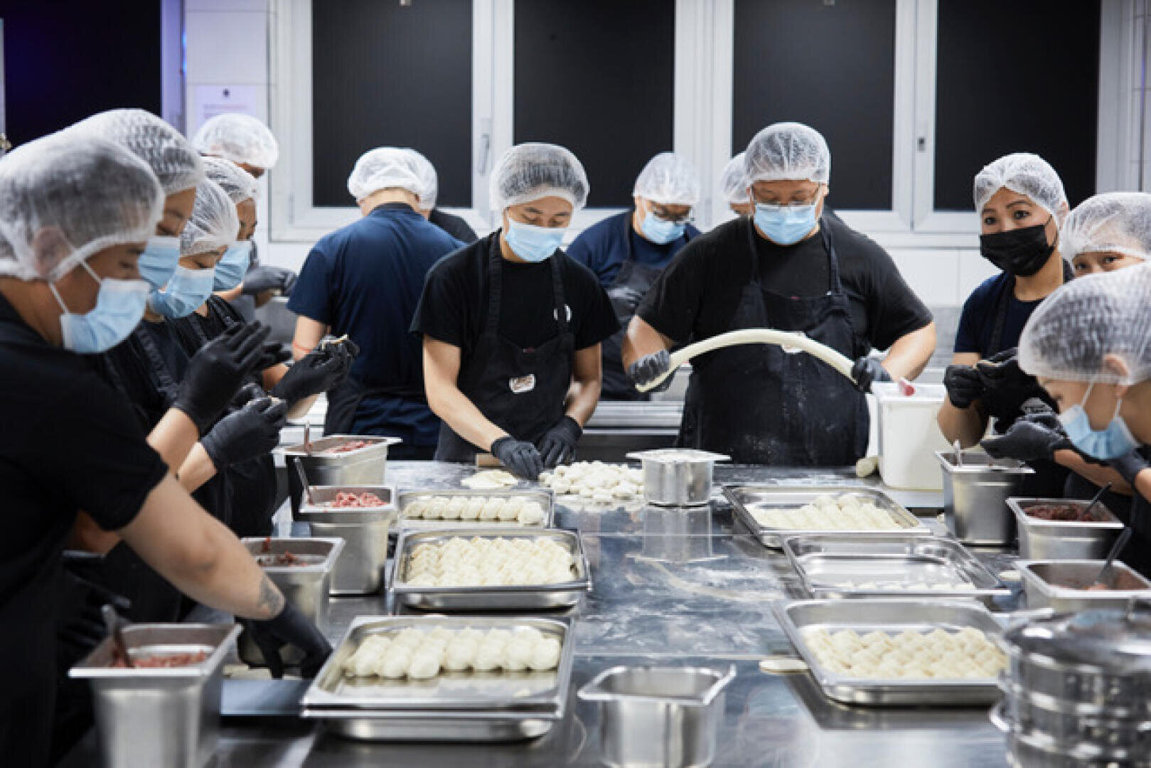 Un groupe de personnes portant des résilles, des masques et des gants travaille ensemble dans une cuisine, préparant et dressant des boulettes sur des plateaux remplis de farine et d'ingrédients.