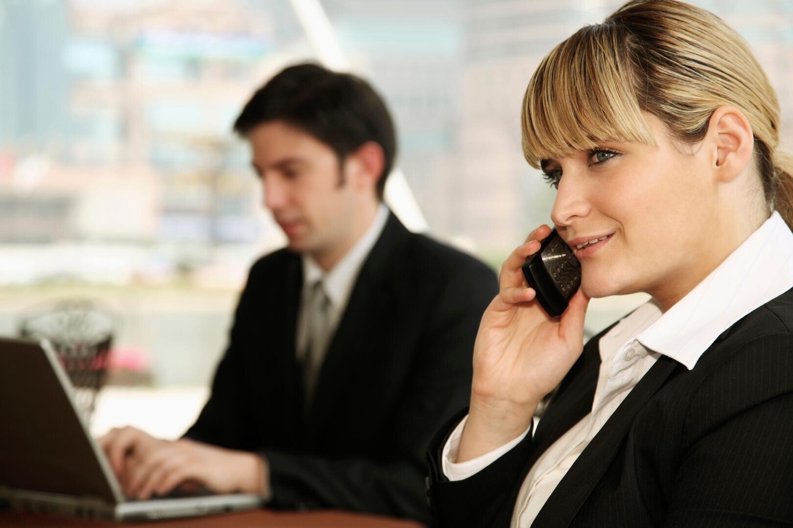 Une femme en costume d'affaires est assise à un bureau et sourit tout en téléphonant. En arrière-plan, un homme en costume travaille sur un ordinateur portable. Tous deux semblent se trouver dans un environnement de bureau moderne.