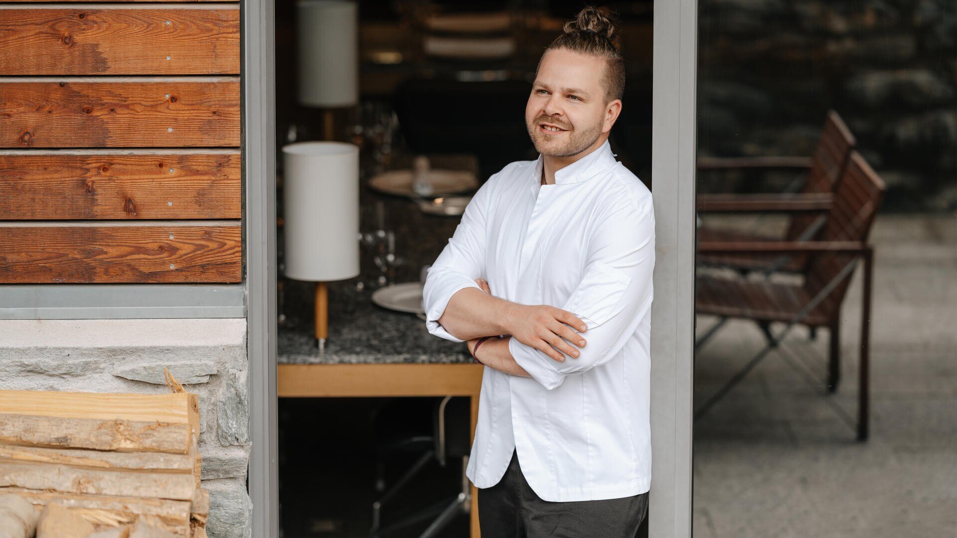 Un cuisinier en blouse blanche se tient souriant, les bras croisés, à l'entrée d'un restaurant moderne avec des détails en bois et en pierre. À l'intérieur, on peut voir des tables et des chaises.