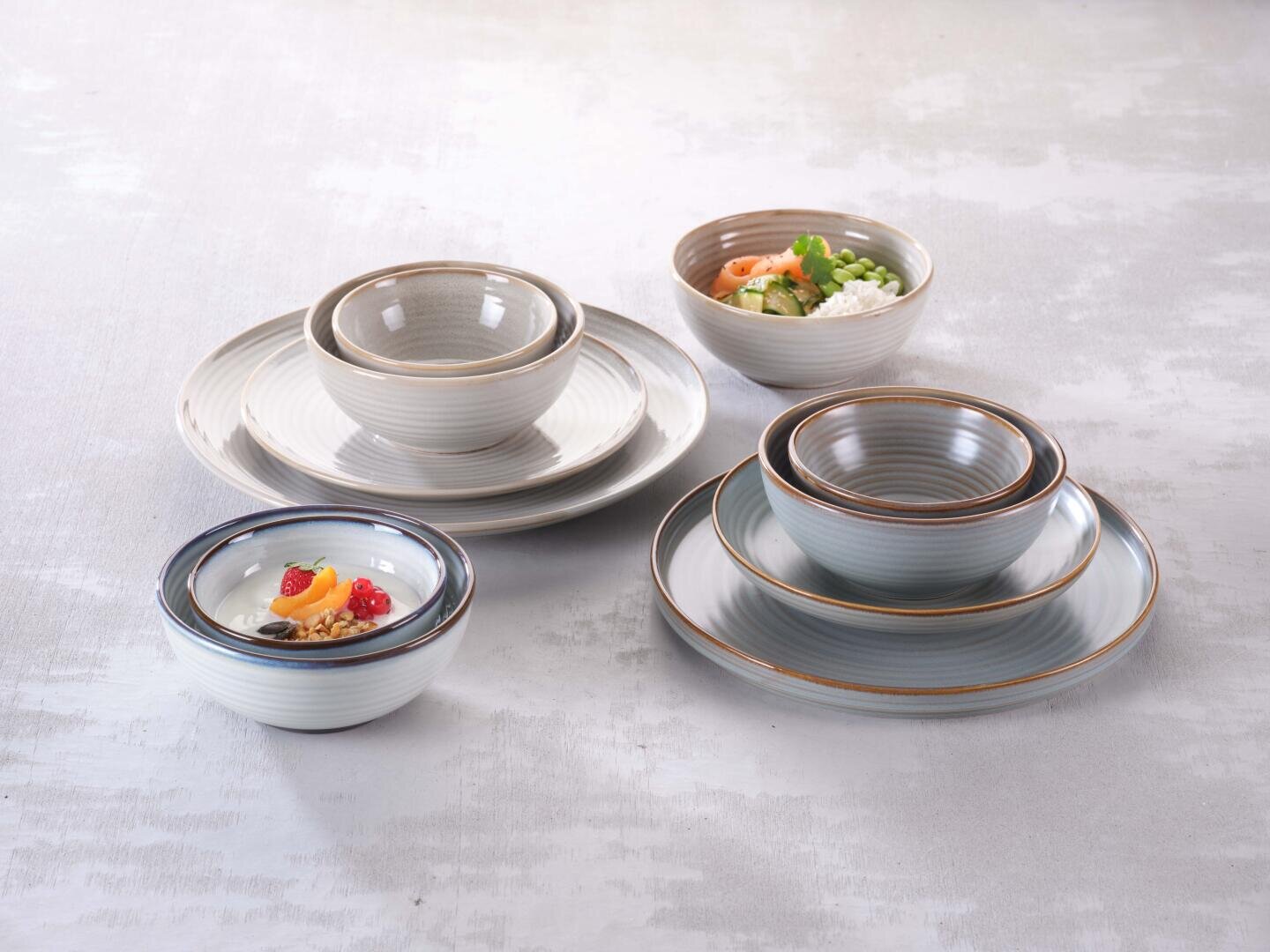 Eine Reihe von Keramiktellern und -schalen in neutralen Tönen, einige gestapelt und einige mit Speisen wie Salat und Dessert, angeordnet auf einer hellgrauen, strukturierten Oberfläche.