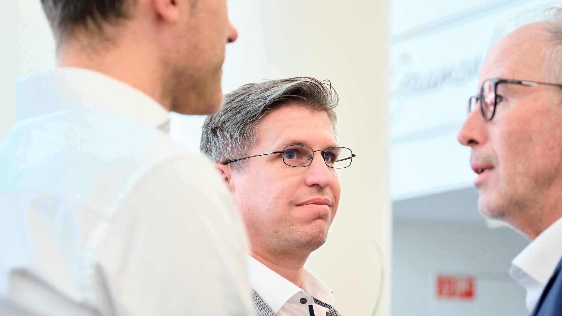 Drei Männer in Geschäftskleidung führen ein Gespräch in einem Haus. Zwei von ihnen sind im Profil dargestellt, während der Mann in der Mitte, der eine Brille trägt, nach vorne schaut und aufmerksam zuhört. Der Hintergrund ist unscharf.