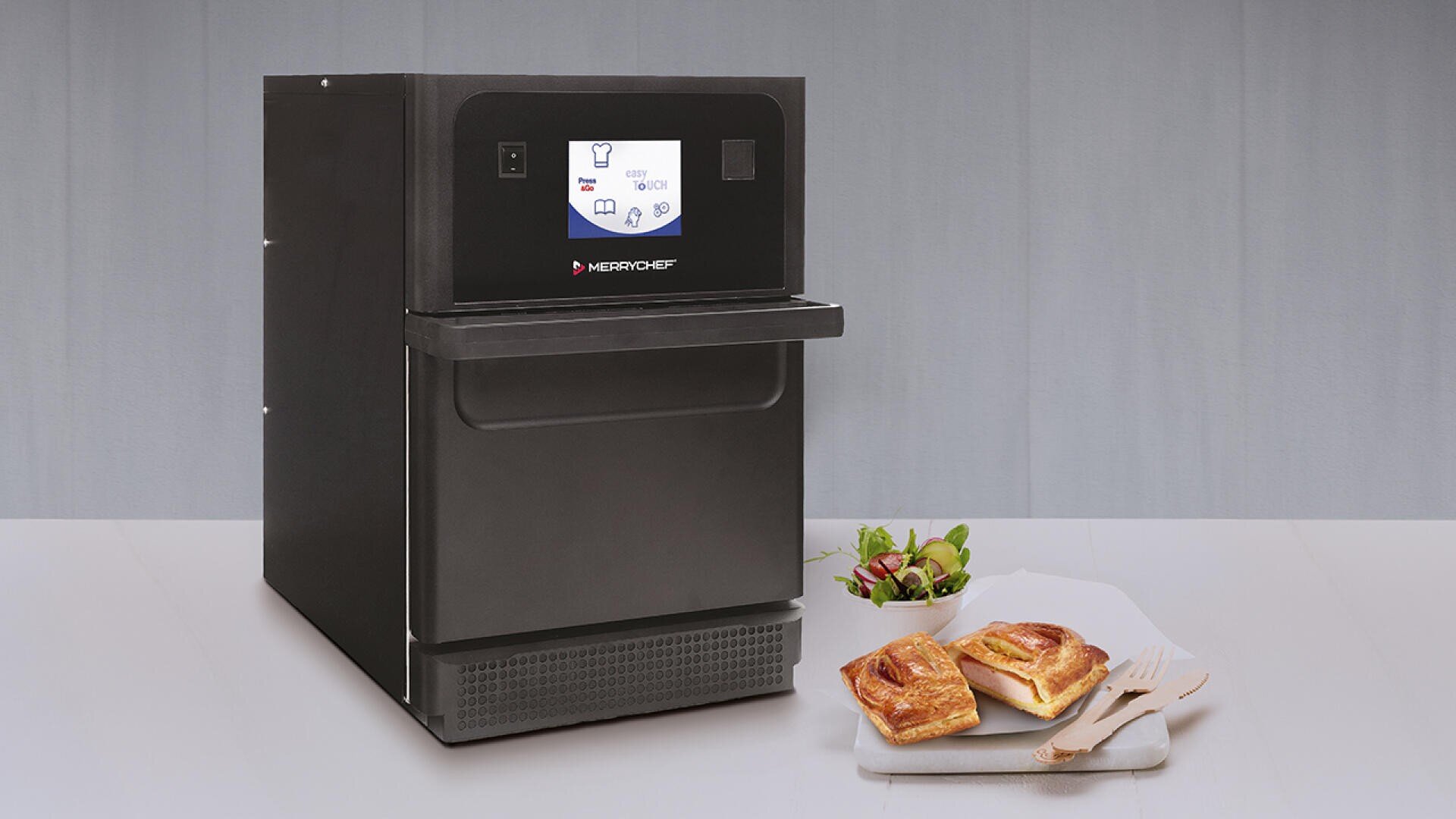 Ein schwarzer kommerzieller Backofen mit Digitalanzeige steht auf einer hellgrauen Oberfläche. Daneben steht ein weißer Teller mit zwei Gebäckstücken, einem kleinen Salat und hölzernen Utensilien. Der Hintergrund ist eine einfarbige hellgraue Wand.