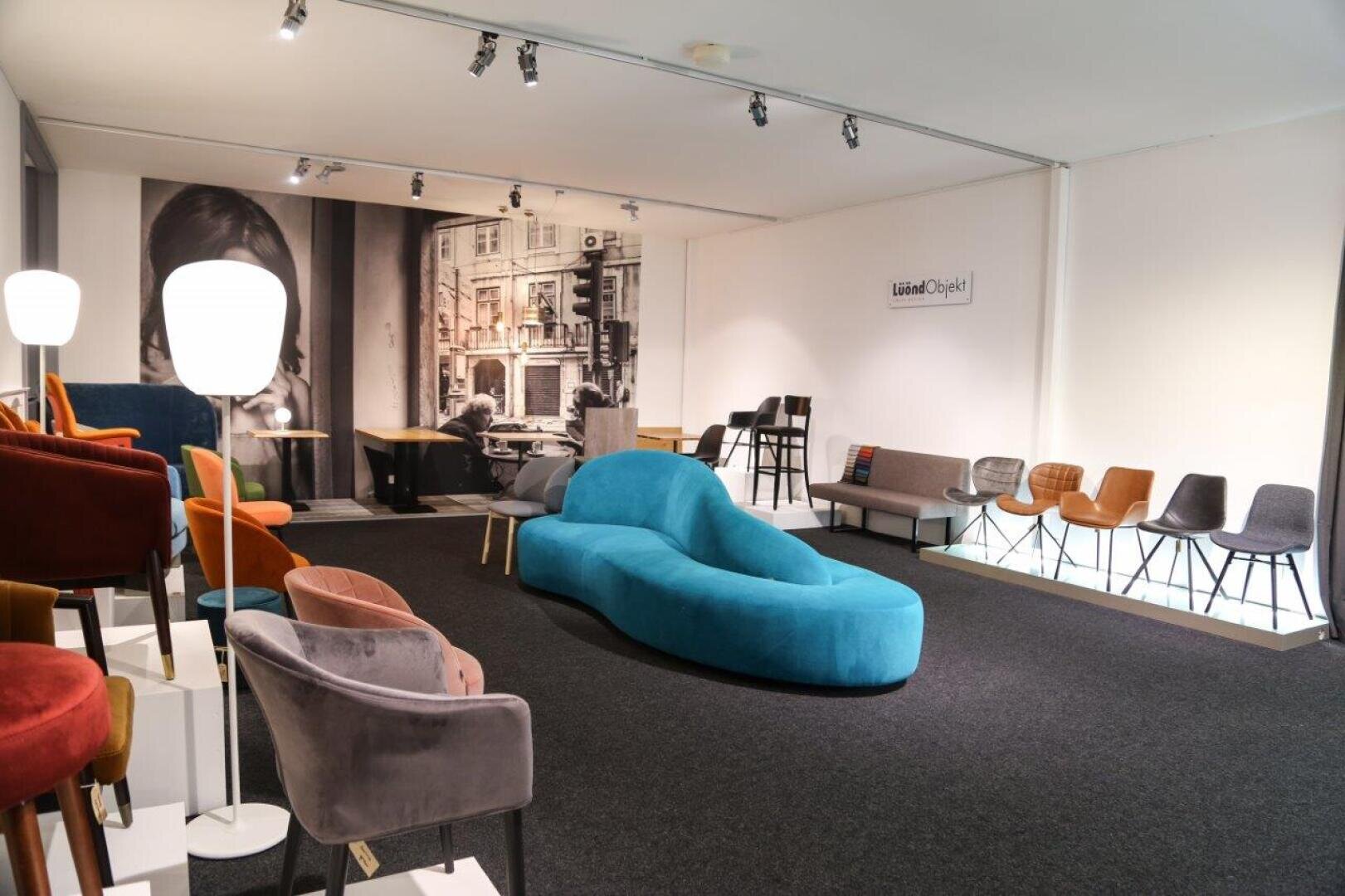 Dans une salle d'exposition moderne, il y a des chaises colorées, un canapé bleu en forme de chaise longue et quelques tables. Une grande fresque murale en noir et blanc représentant des vues de la ville est accrochée aux murs, ainsi qu'un panneau portant l'inscription Listed Design. La pièce est bien éclairée.