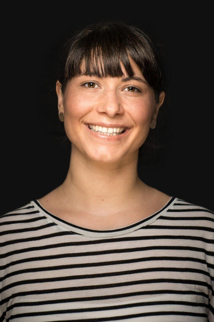 Eine Frau mit dunklem Haar und Pony lächelt in die Kamera. Sie trägt ein schwarz-weiß gestreiftes Hemd und posiert vor einem schwarzen Hintergrund.