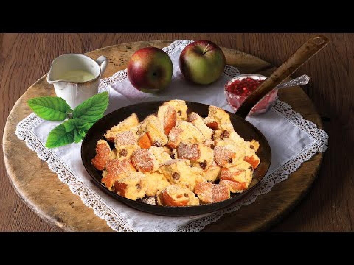 Auf einem Holzbrett steht eine Pfanne mit Kaiserschmarrn-Stückchen, die mit Puderzucker bestreut sind, daneben zwei Äpfel, ein Minzezweig, ein kleiner Krug mit Sahne und ein Teller mit roter Marmelade.