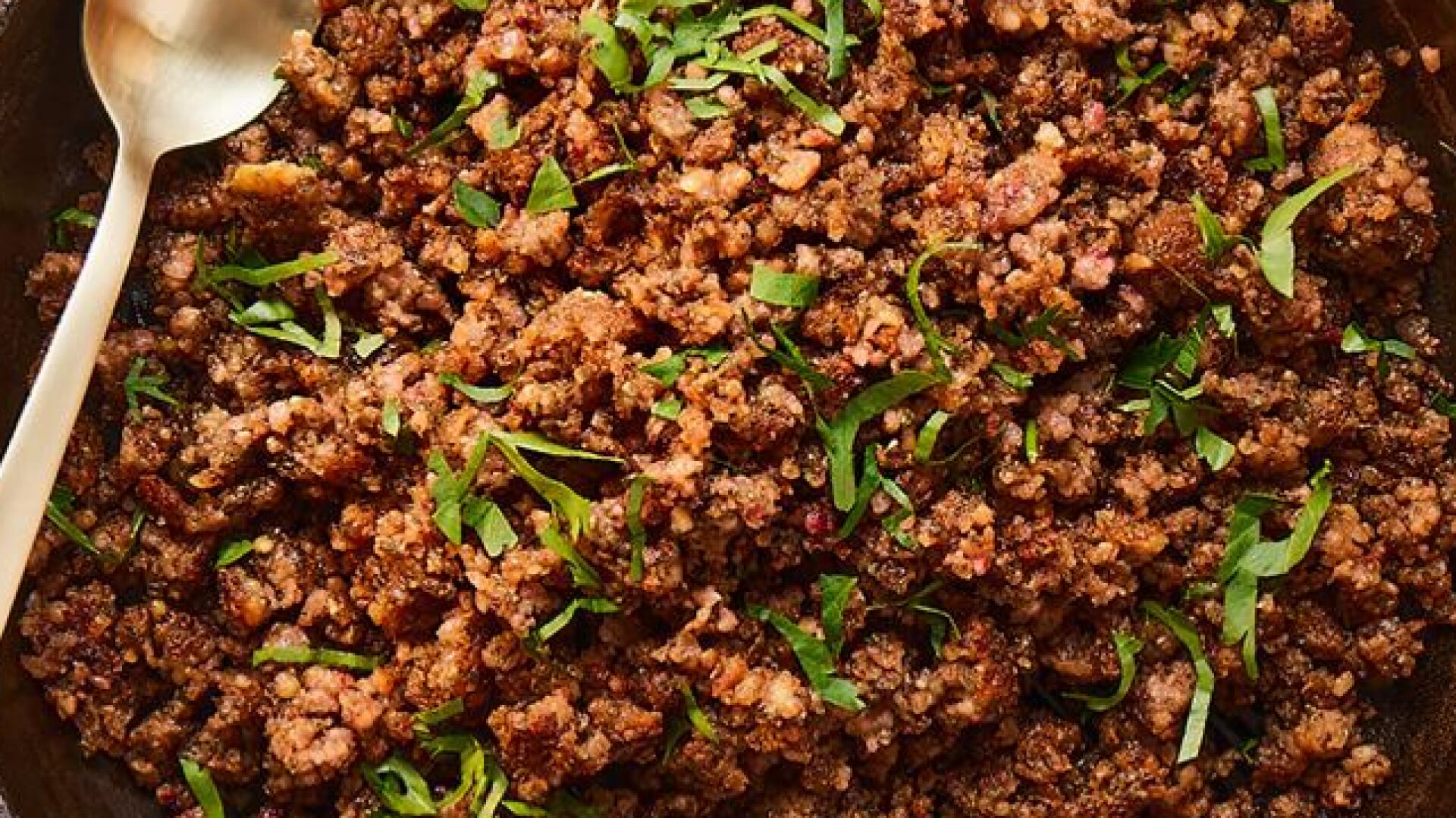 Eine Pfanne mit gekochtem, gewürztem veganen Hack aus Mungbohne und gehackten frischen Kräutern, wobei ein goldener Löffel auf dem Pfannenrand liegt.