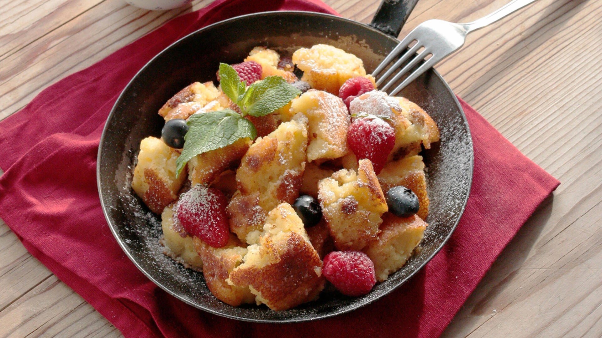 Eine Pfanne mit goldgelben, fluffigen Pfannkuchenstücken, die mit Puderzucker, Himbeeren, Blaubeeren und einem Minzzweig belegt sind, steht auf einer roten Stoffserviette auf einem Holztisch, auf dem eine Gabel liegt.