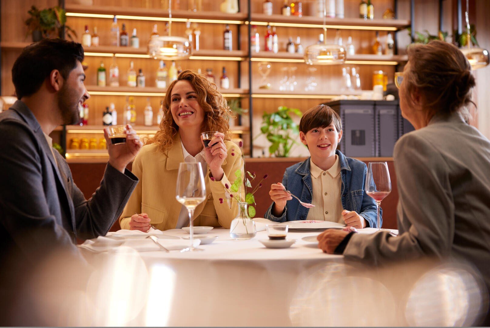 Vier Personen, darunter ein Kind, sitzen an einem runden Restauranttisch mit Getränken und Tellern, lächeln und unterhalten sich. Hinter ihnen stehen Regale mit Flaschen und Pflanzen, die eine warme, einladende Atmosphäre schaffen.