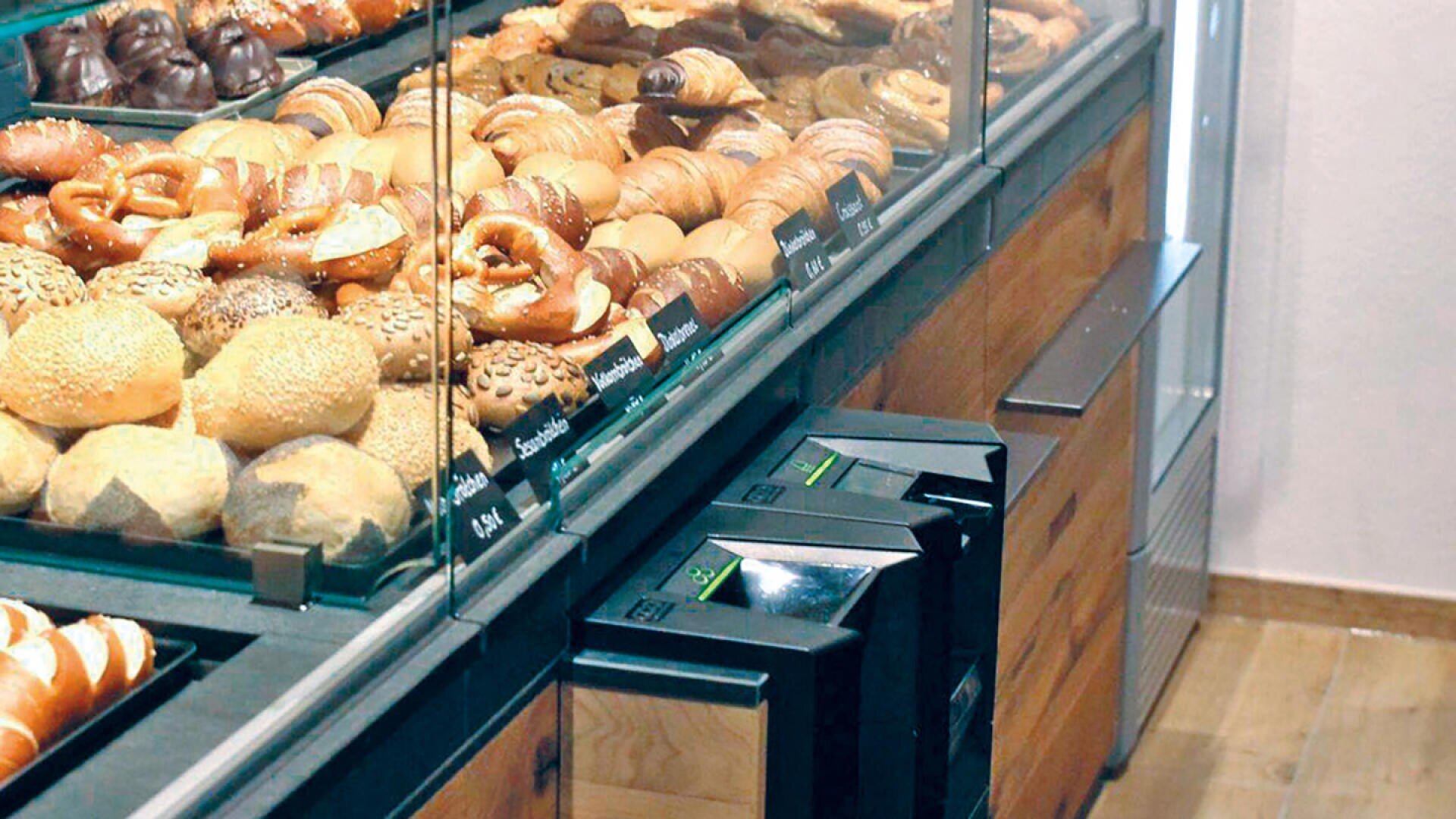 Eine Bäckereivitrine, gefüllt mit verschiedenen Brotsorten und Gebäck, darunter Brötchen, Croissants und Brezeln, mit Preisschildern auf dem Glas und zwei schwarzen Verkaufsautomaten auf der Holztheke darunter.