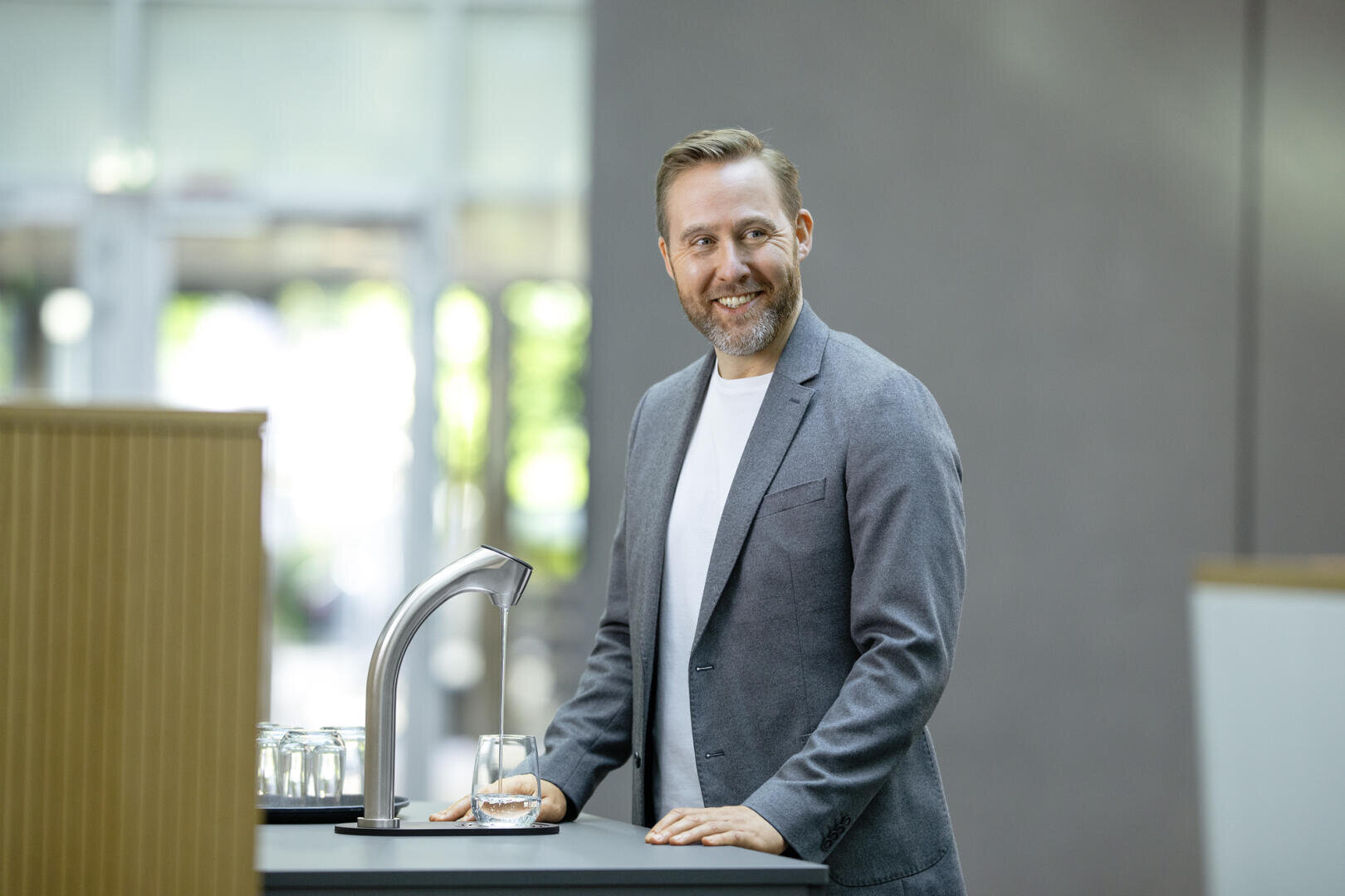 Un homme en blazer gris et chemise blanche sourit, debout à une table, remplissant un verre d'eau d'une fontaine moderne dans un intérieur lumineux.