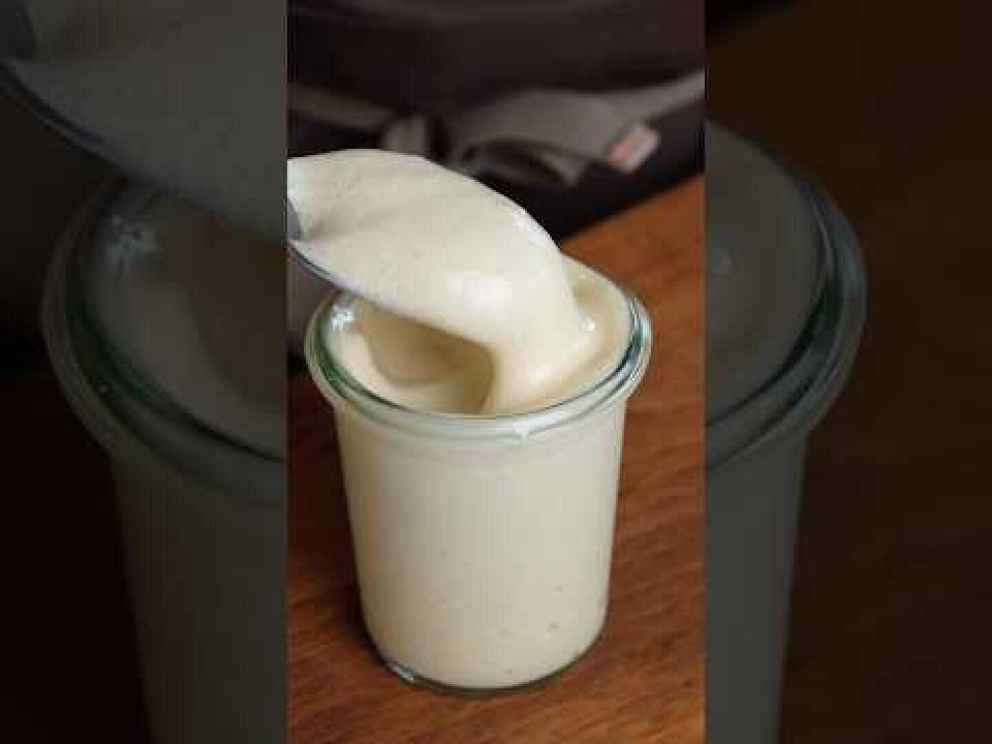 Ein Glas mit einer dicken, cremigen, hellgelben Soße oder einem Aufstrich steht auf einer Holzoberfläche. Mit einem Löffel wird etwas von der glatten Mischung herausgenommen.