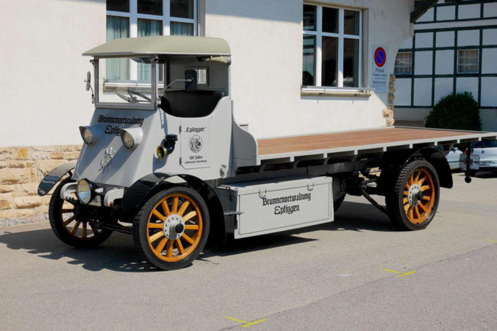 Ein alter Pritschenwagen mit Holzspeichenrädern und einem grauen Aufbau ist auf einer Straße vor einem Gebäude geparkt. Der Lkw hat ein offenes Fahrerhaus und eine deutsche Beschriftung an der Seite.