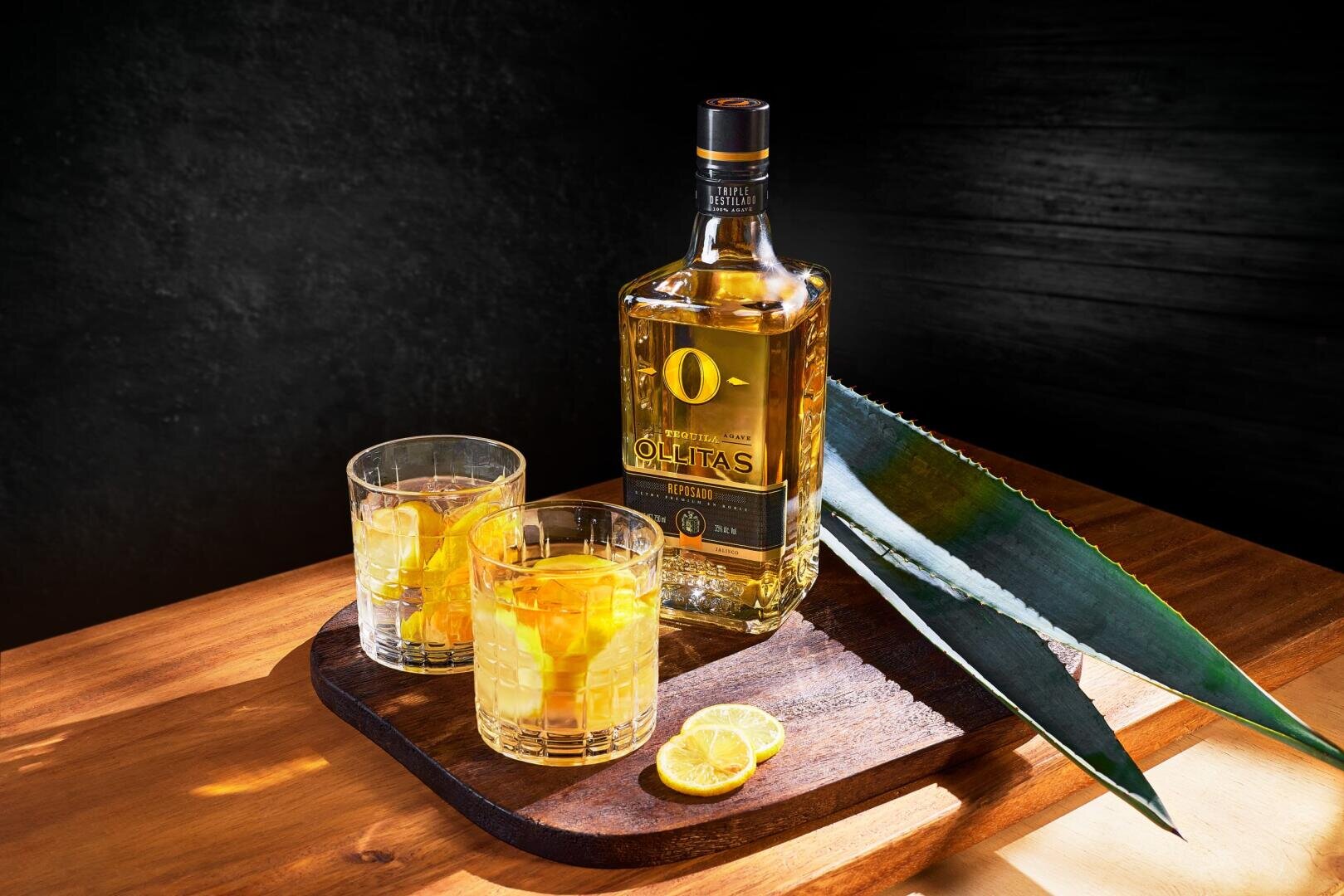Eine Flasche Olitas Tequila steht auf einem Holztablett mit zwei Cocktailgläsern, Eiswürfeln, Agavenblättern und Zitronenscheiben auf einer hölzernen Oberfläche vor einem dunklen Hintergrund.