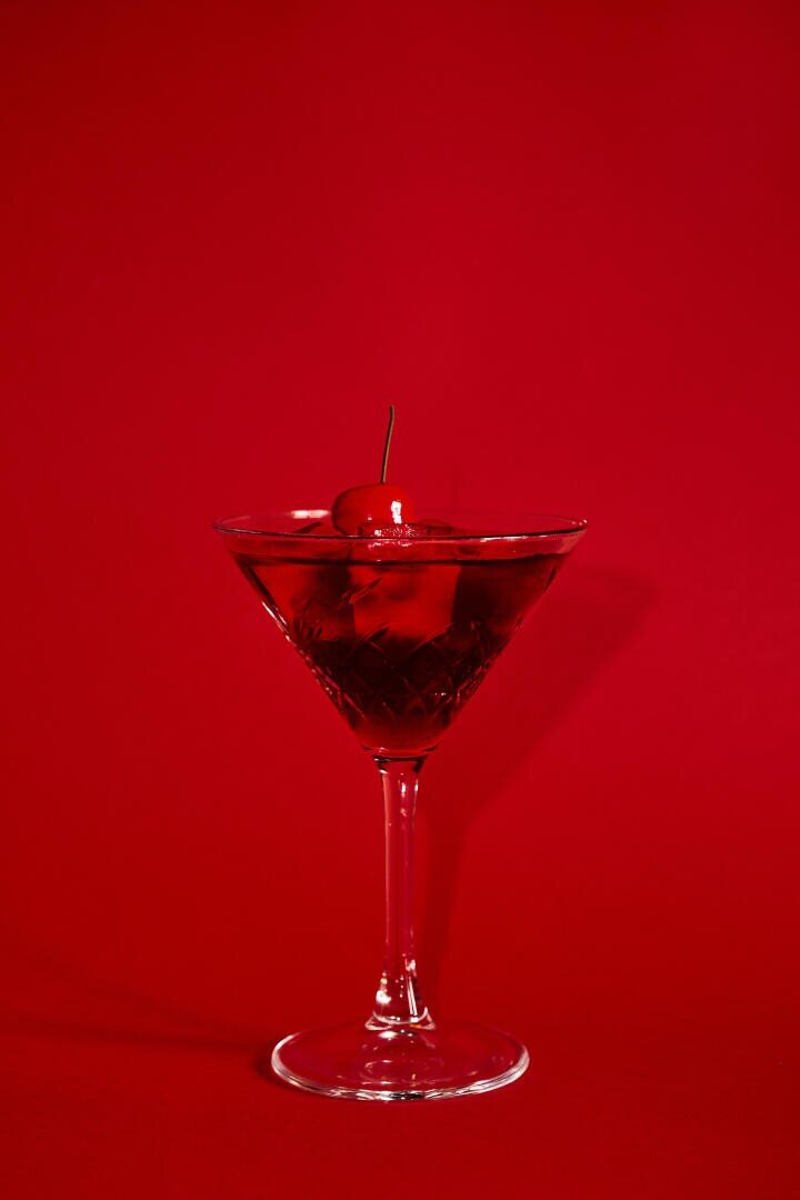 Ein klares Martini-Glas, gefüllt mit einem roten Cocktail und Eis, garniert mit einer Kirsche auf der Spitze, vor einem einfarbig roten Hintergrund.