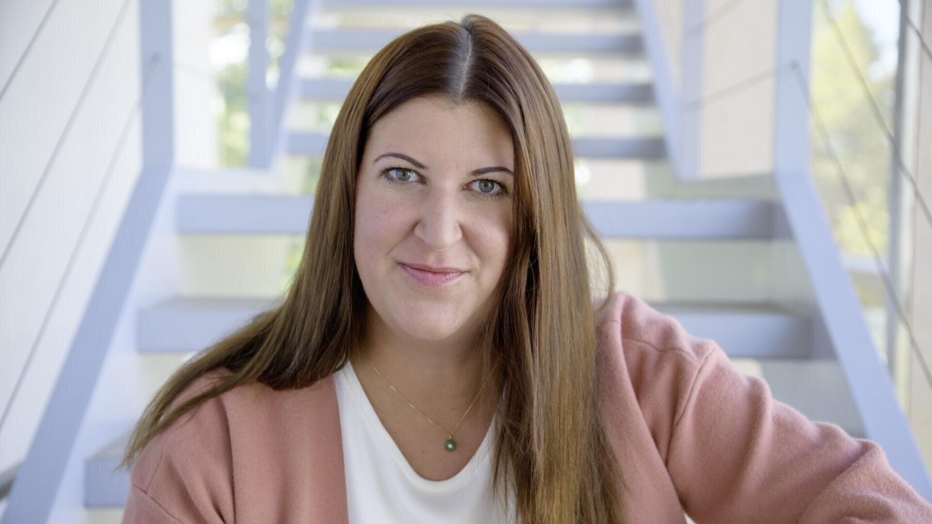 Eine Frau mit langen braunen Haaren, die eine rosa Strickjacke und ein weißes Oberteil trägt, sitzt sanft lächelnd vor einer modernen Treppe mit Glasgeländern.