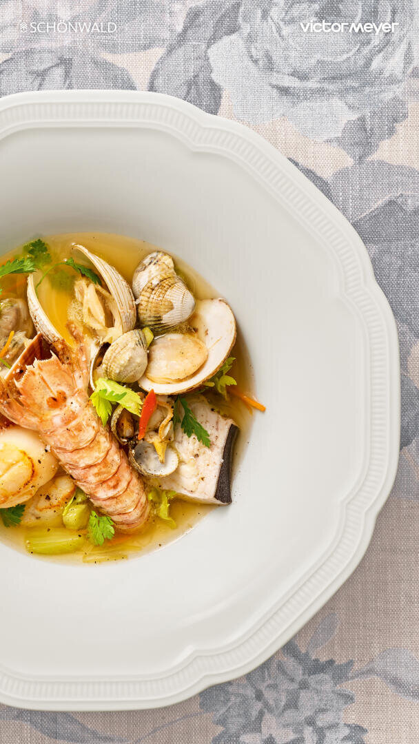 Eine weiße Schale enthält einen Meeresfrüchteeintopf mit Hummerschwanz, Venusmuscheln, Fischstücken, Gemüse und Kräutern in einer klaren Brühe, die auf einem gemusterten Tischtuch platziert ist.