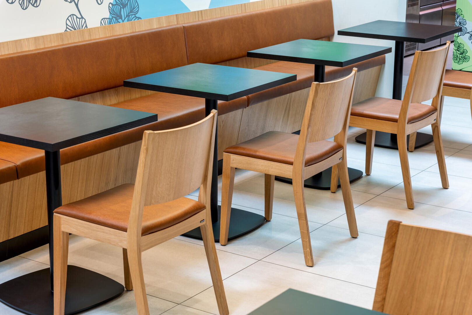 Une rangée de chaises en bois vides et de tables carrées noires à côté d'une banquette marron rembourrée dans un café moderne et bien éclairé au sol carrelé et à l'art mural décoratif.