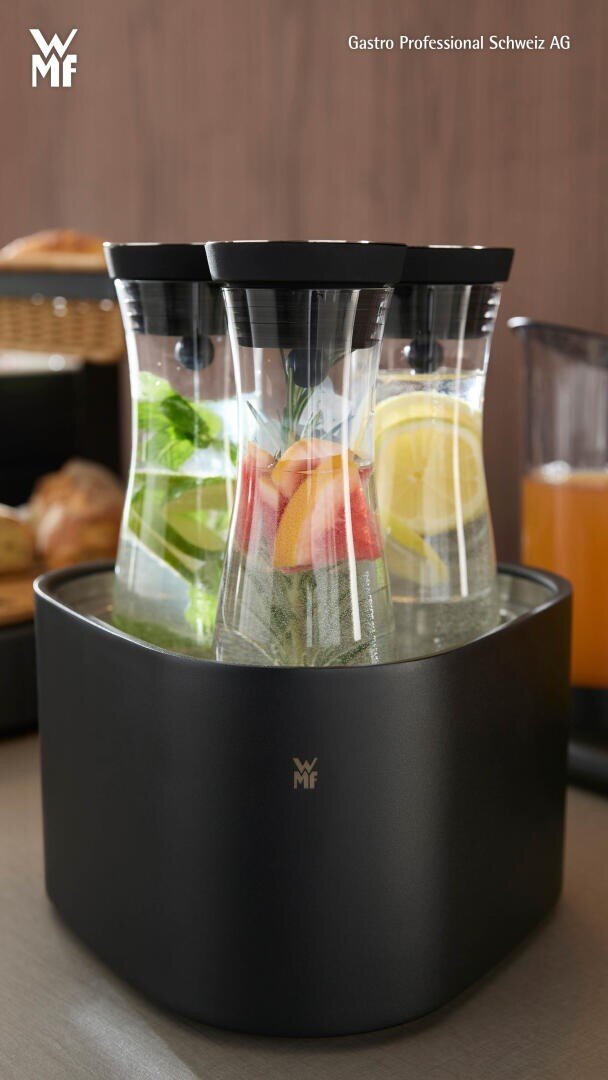 Trois carafes en verre remplies d'eau enrichie d'agrumes, de menthe et de rondelles de concombre sont posées sur un comptoir dans un élégant refroidisseur de boissons WMF noir, avec des pâtisseries et une boisson à l'orange en arrière-plan.