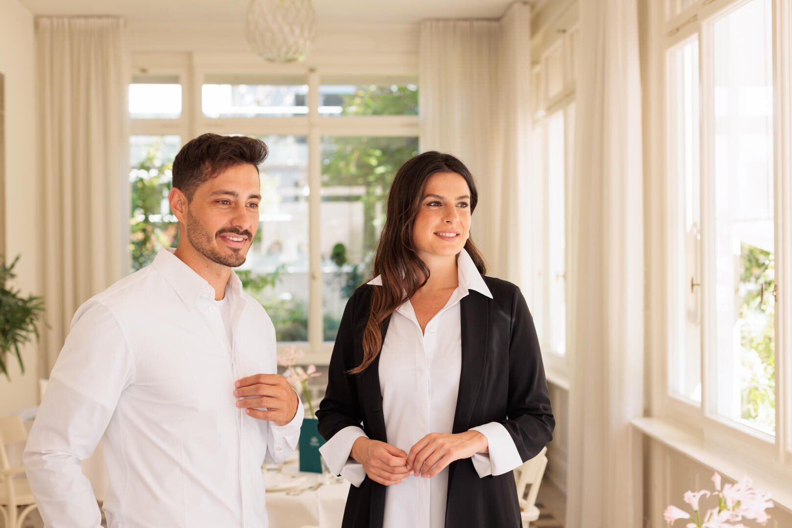 Ein Mann in einem weißen Hemd und eine Frau in einem schwarzen Blazer stehen zusammen in einem Innenraum, lächeln und schauen aus einem großen Fenster in einem hellen, eleganten Raum mit weißen Vorhängen und natürlichem Licht.
