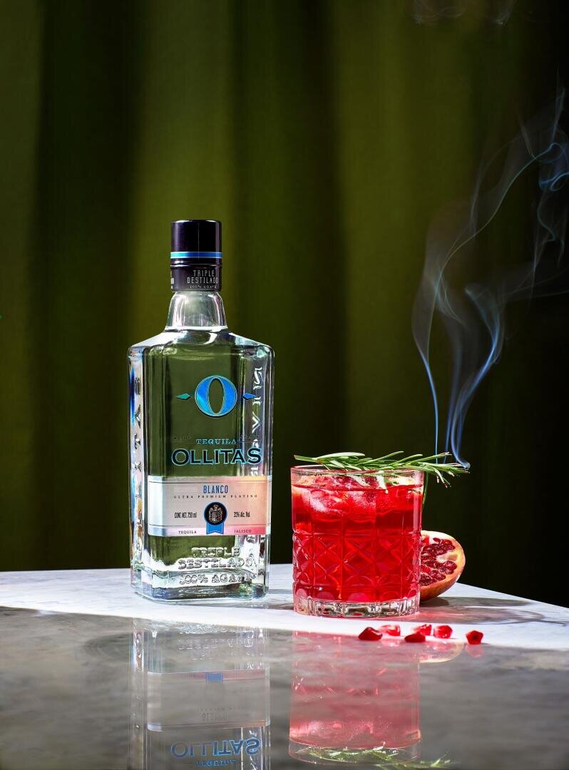 Eine Flasche Ollitas Blanco Tequila steht neben einem roten Cocktail, der mit Rosmarin und Granatapfelkernen garniert ist, wobei Rauch aus der Garnitur aufsteigt. Ein halbierter Granatapfel steht im Hintergrund auf einer Marmorplatte.