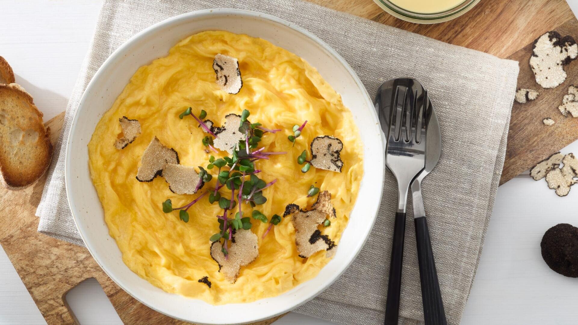 Un bol d'œufs brouillés crémeux garnis de lamelles de truffe noire et de microgreens, posés sur une serviette beige, avec une fourchette et une cuillère à côté. Du pain grillé et des lamelles de truffe supplémentaires se trouvent à proximité sur une planche en bois.