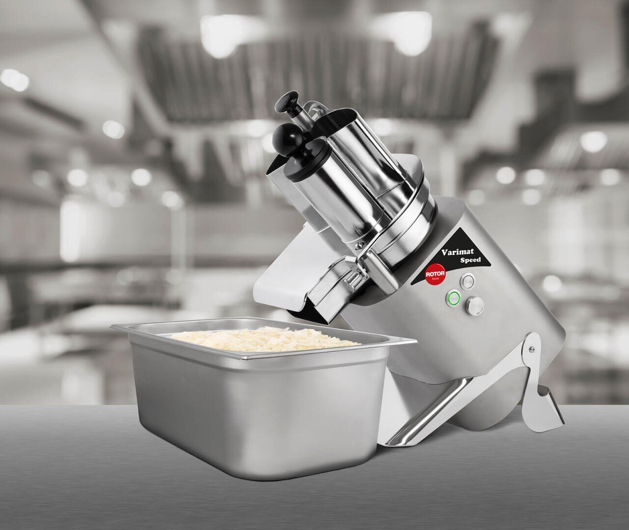 Eine handelsübliche Küchenmaschine steht auf einem Tresen in einer Küche, hinter einem Metallbehälter, der mit geriebenem Käse gefüllt ist. Der Hintergrund ist leicht unscharf und zeigt Küchengeräte und Lichter.