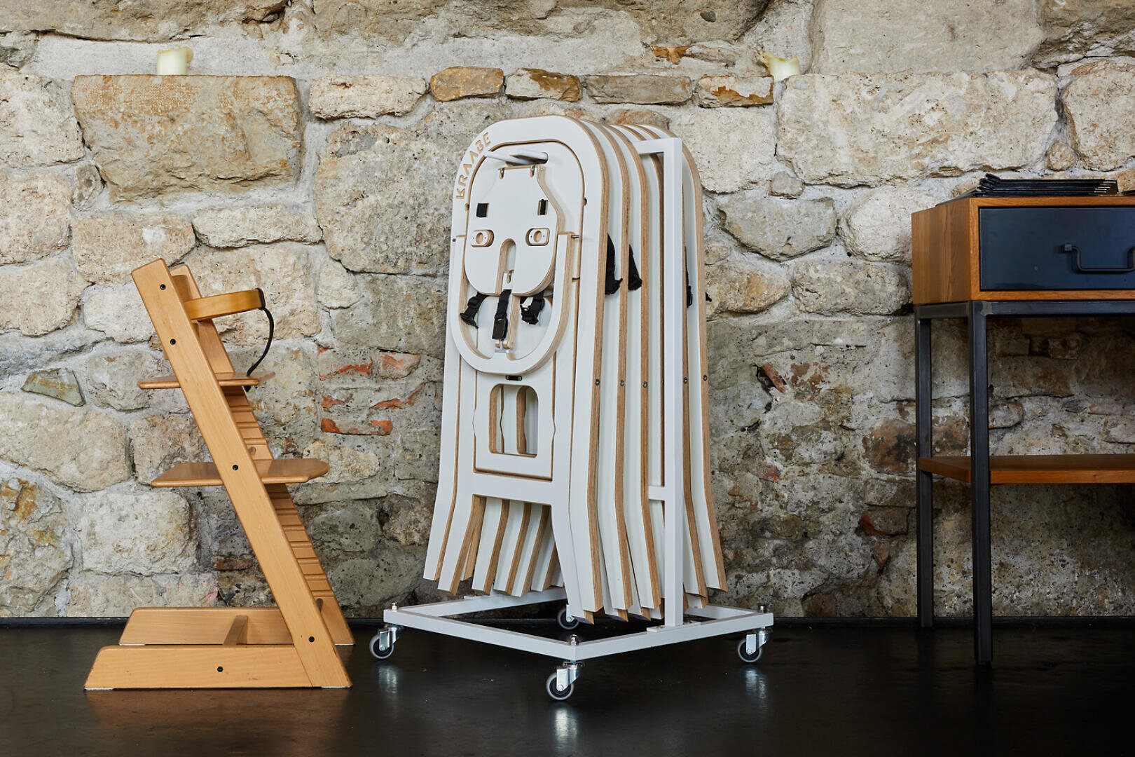 Ein hölzerner Hochstuhl steht neben mehreren gestapelten weißen Hochstühlen auf einem Gestell auf Rädern an einer rustikalen Steinwand neben einem kleinen Holztisch mit einer schwarzen Schublade.