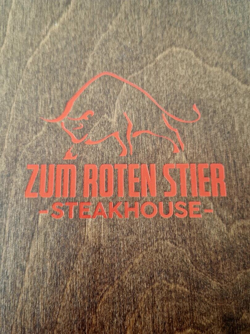 Rote Strichzeichnung eines Stiers über der Aufschrift "ZUM ROTEN STIER - STEAKHOUSE -" in fetten roten Buchstaben auf dunklem Holzuntergrund.