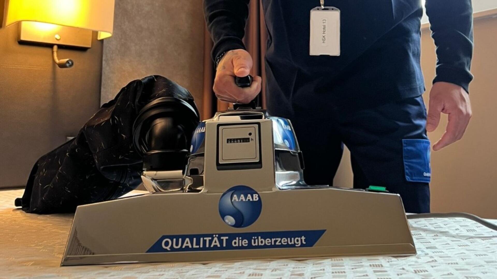 Eine dunkel gekleidete Person bedient eine große, glänzende Metallmaschine mit der Aufschrift AAAB QUALITÄT die überzeugt auf einem Bett mit einem gemusterten Bezug, neben einer schwarzen Tasche in einem Raum mit gedämpfter Beleuchtung.