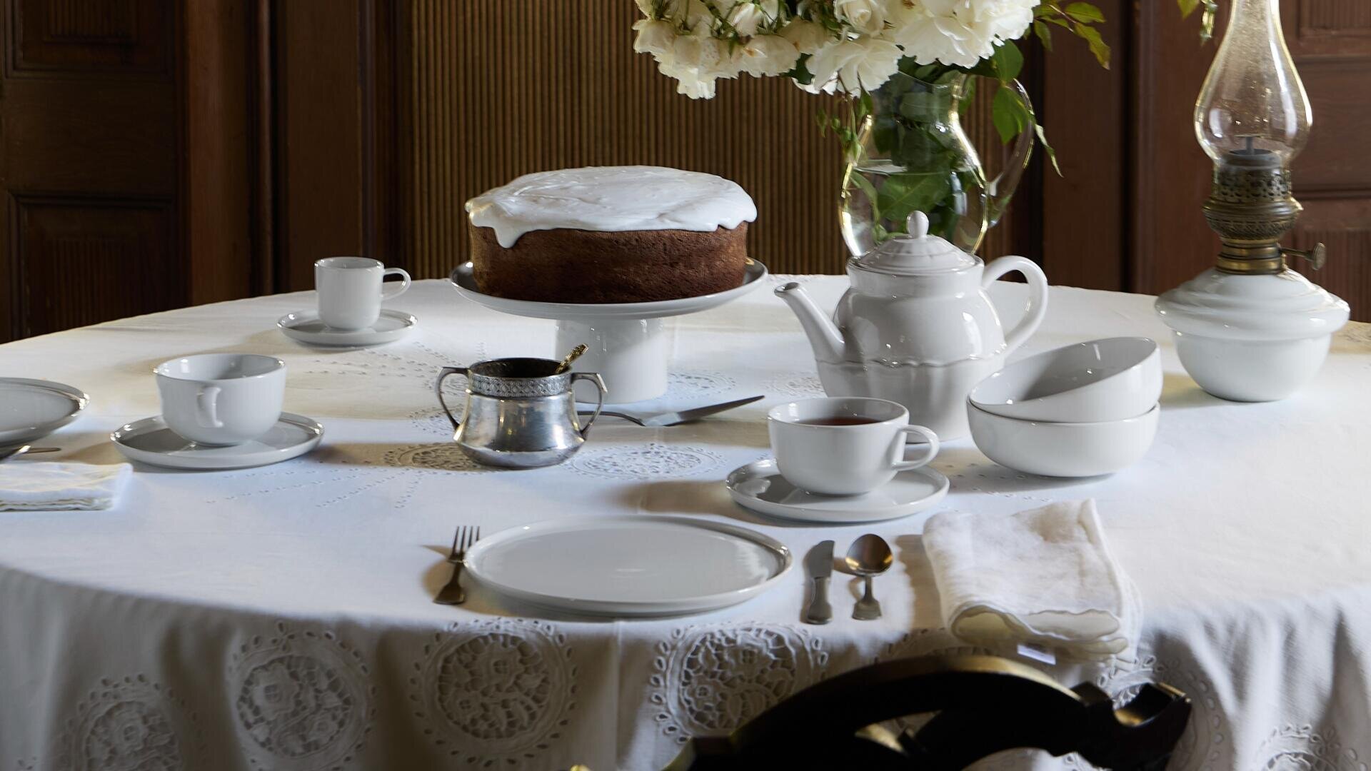 Ein runder, für den Tee gedeckter Tisch mit weißem Porzellan, einer Teekanne, Tassen, Tellern, Besteck, einem großen Eiskuchen auf einem Ständer, einem Milchkännchen und einer Vase mit weißen Blumen auf einer weißen Spitzentischdecke.