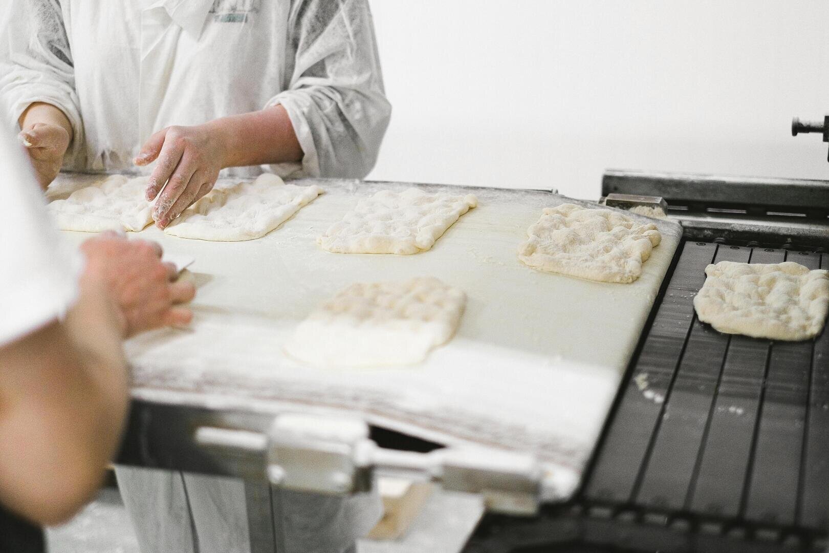 Deux personnes en uniforme blanc forment et pressent des morceaux de pâte sur une table enfarinée dans une boulangerie ou une cuisine et les préparent pour la cuisson. Plusieurs morceaux de pâte rectangulaires sont visibles sur la surface.