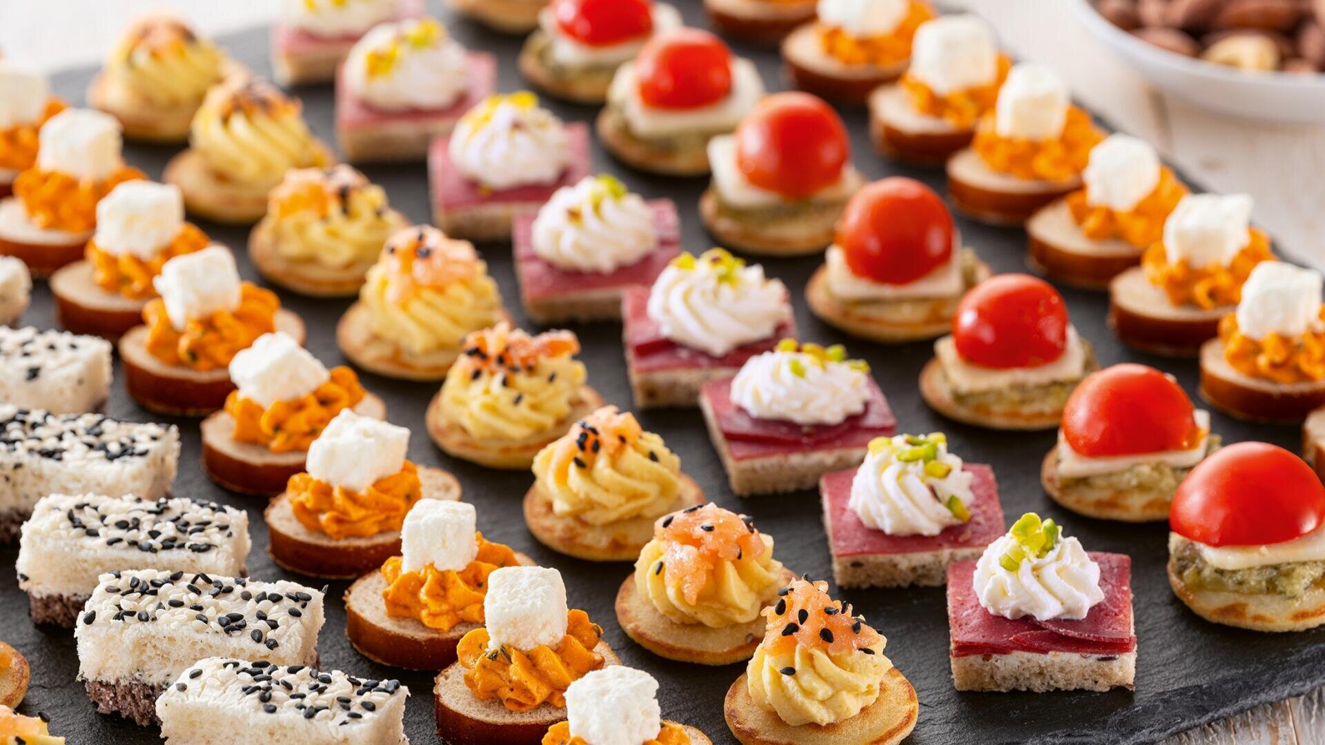 Ein Sortiment bunter, mundgerechter Häppchen auf einer schwarzen Schieferplatte, darunter Canapés mit Sahne, Kirschtomaten, Schlagsahne, Kräutern, Samen, Käsewürfeln und Brotscheiben mit verschiedenen Garnierungen.
