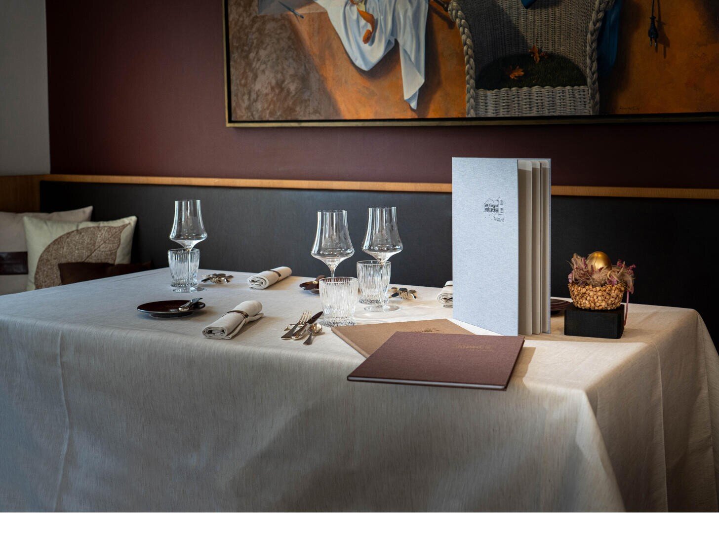 Ein ordentlich gedeckter Restauranttisch mit weißem Tischtuch, Gläsern, Besteck, Servietten, einer Speisekarte und einem kleinen dekorativen Tafelaufsatz; im Hintergrund hängt ein Gemälde an der Wand.