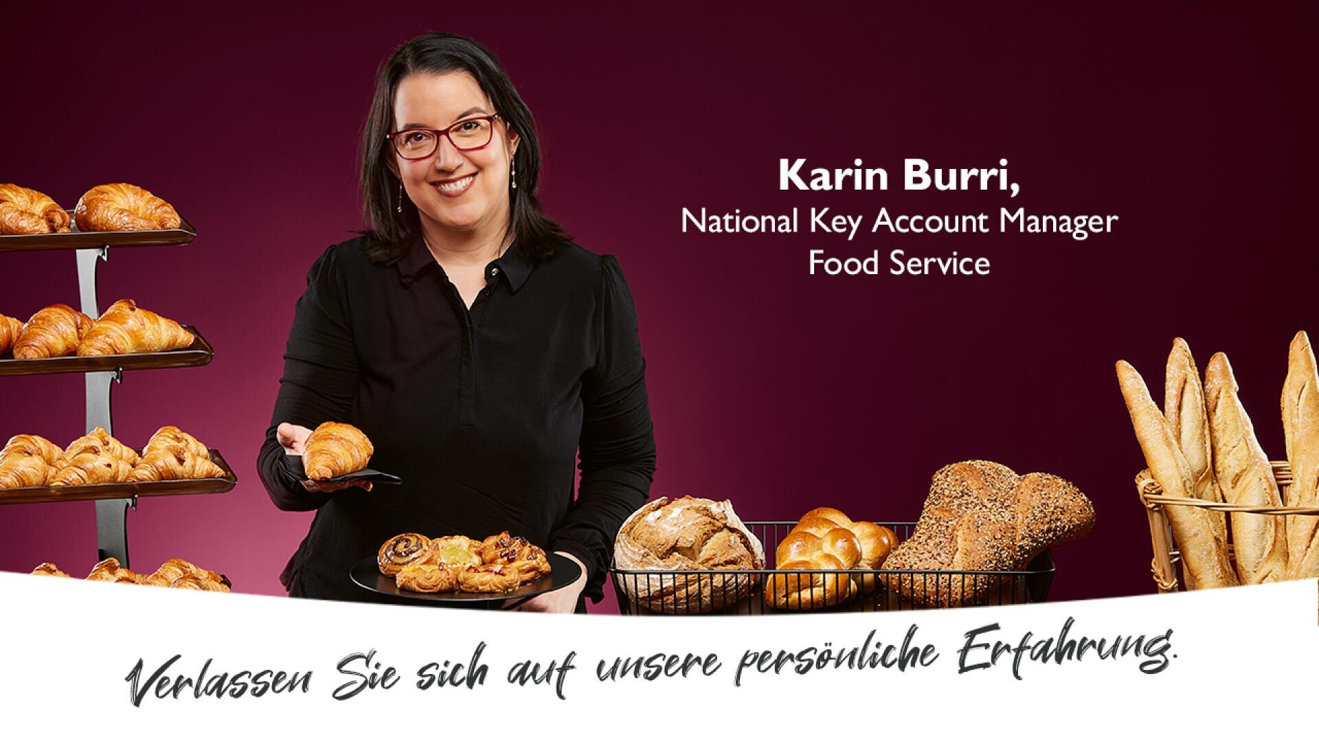 Eine Frau mit Brille und schwarzer Kleidung hält ein Tablett mit Gebäck und steht neben Körben und Tabletts mit Croissants, Brötchen und Baguettes. Der Text stellt sie als Karin Burri vor, National Key Account Manager Food Service.