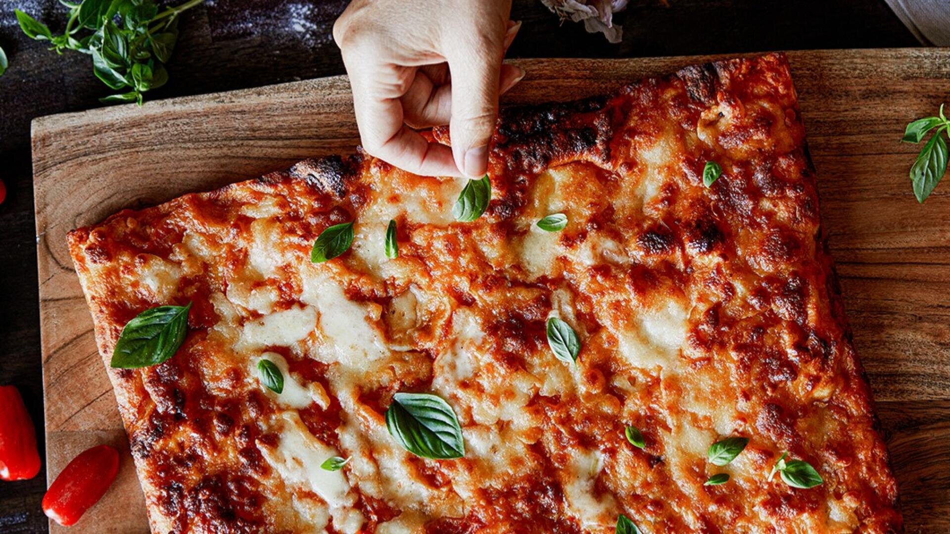 Eine Hand legt ein frisches Basilikumblatt auf eine rechteckige Pizza mit geschmolzenem Käse und Tomatensauce, die auf einem Holzbrett liegt.