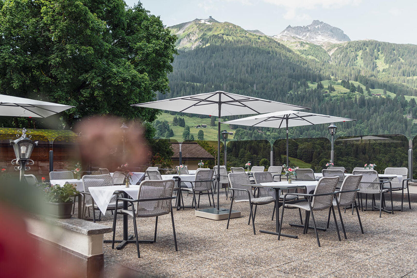 Terrasse mit Tischen und Stühlen unter großen Sonnenschirmen, mit Blick auf grüne Hügel, Wälder und ferne Berge unter einem teilweise bewölkten Himmel. Blumen und ein Baum tragen zur friedlichen Atmosphäre bei.