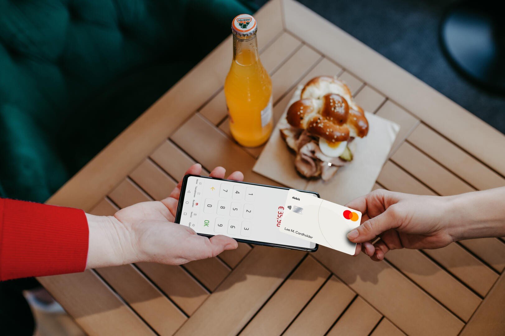 Une personne paie avec une carte de crédit Mastercard tapée sur un smartphone tenu par une autre personne. Sur la table se trouvent une bouteille de boisson à l'orange et un sandwich avec de la sauce, posés sur une serviette.