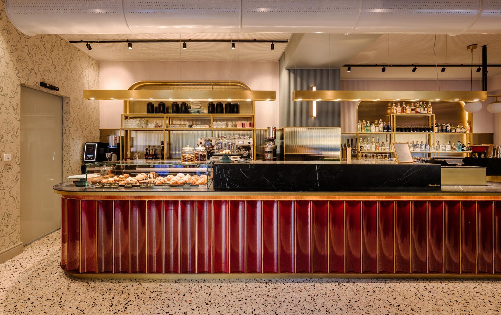 Un comptoir de boulangerie moderne avec un plateau en marbre noir et des panneaux avant rouges présente des pâtisseries à l'intérieur d'une vitrine. Derrière le comptoir se trouvent des étagères avec des bocaux, une machine à café et des bouteilles, le tout dans un intérieur élégant et chaleureusement éclairé.