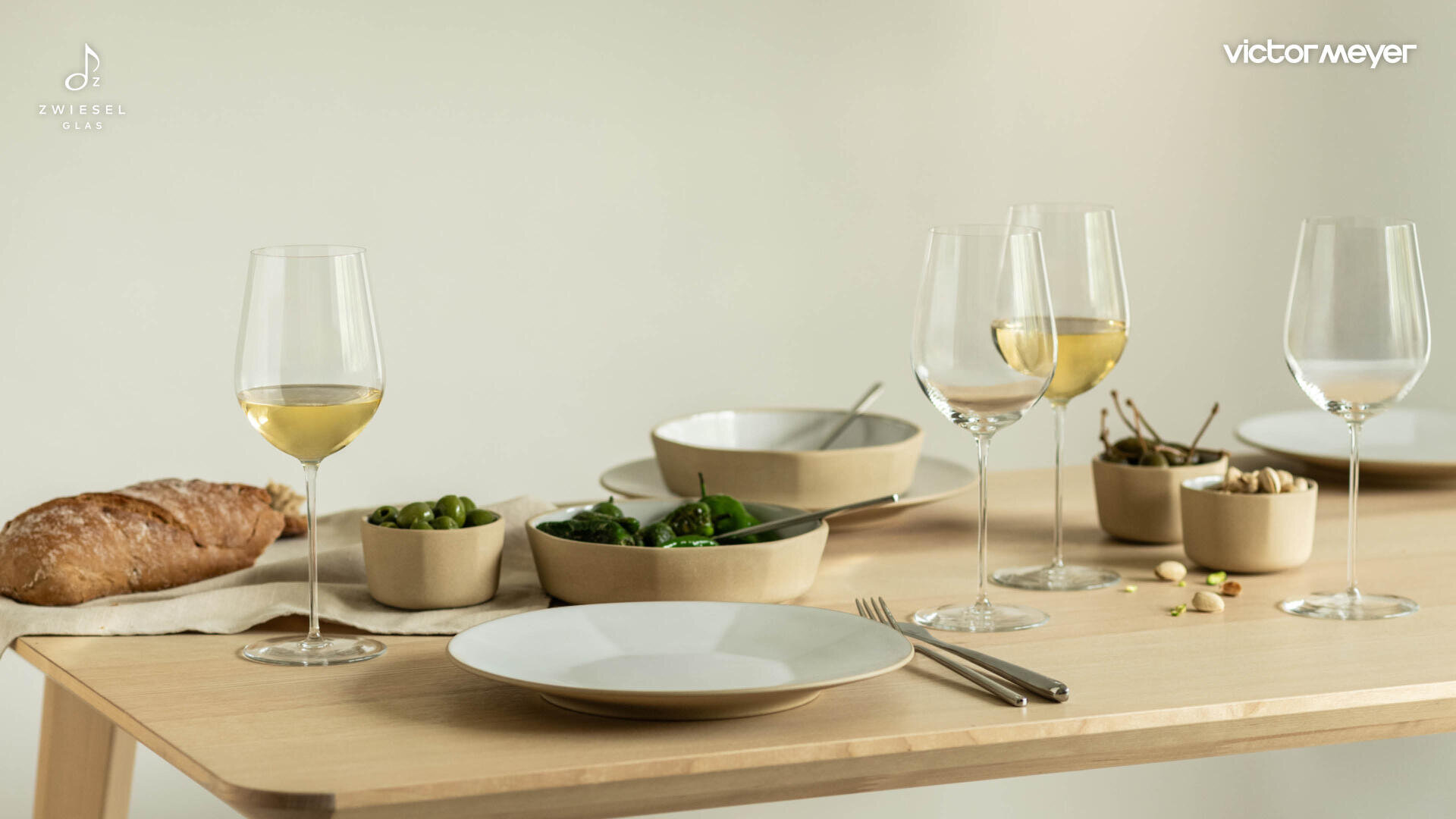 Ein minimalistisch gedeckter Tisch mit weißen Tellern, Weingläsern mit Weißwein, Schalen mit grünem Gemüse, Nüssen und einem Laib Brot auf einem hellen Holztisch vor einem schlichten Hintergrund.