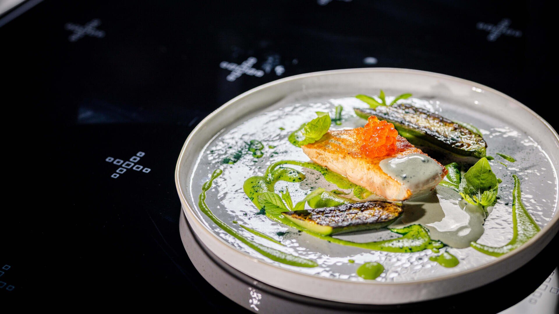Ein Gourmetgericht aus einem Stück angebratenem Fisch, garniert mit Orangeneiern, dazu zwei Stücke angebratene Makrele, eine grüne Soße, frische Kräuter und eine cremige weiße Soße auf einem weißen runden Teller.