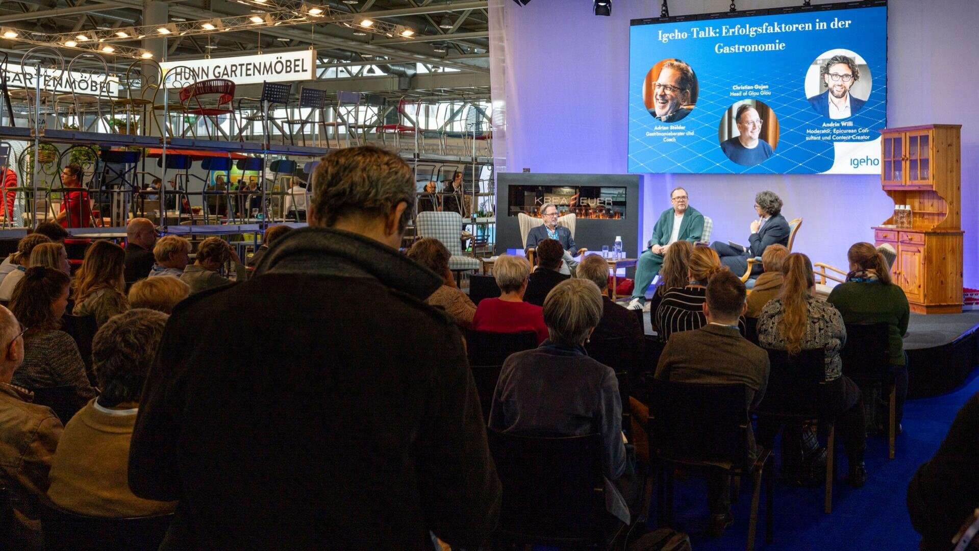 Ein Publikum verfolgt eine Podiumsdiskussion auf einer Fachmesse. Drei Redner sitzen auf der Bühne, ihre Fotos und Namen werden auf einem großen Bildschirm darüber angezeigt. Im Hintergrund sind Messestände und Gartenmöbel zu sehen.