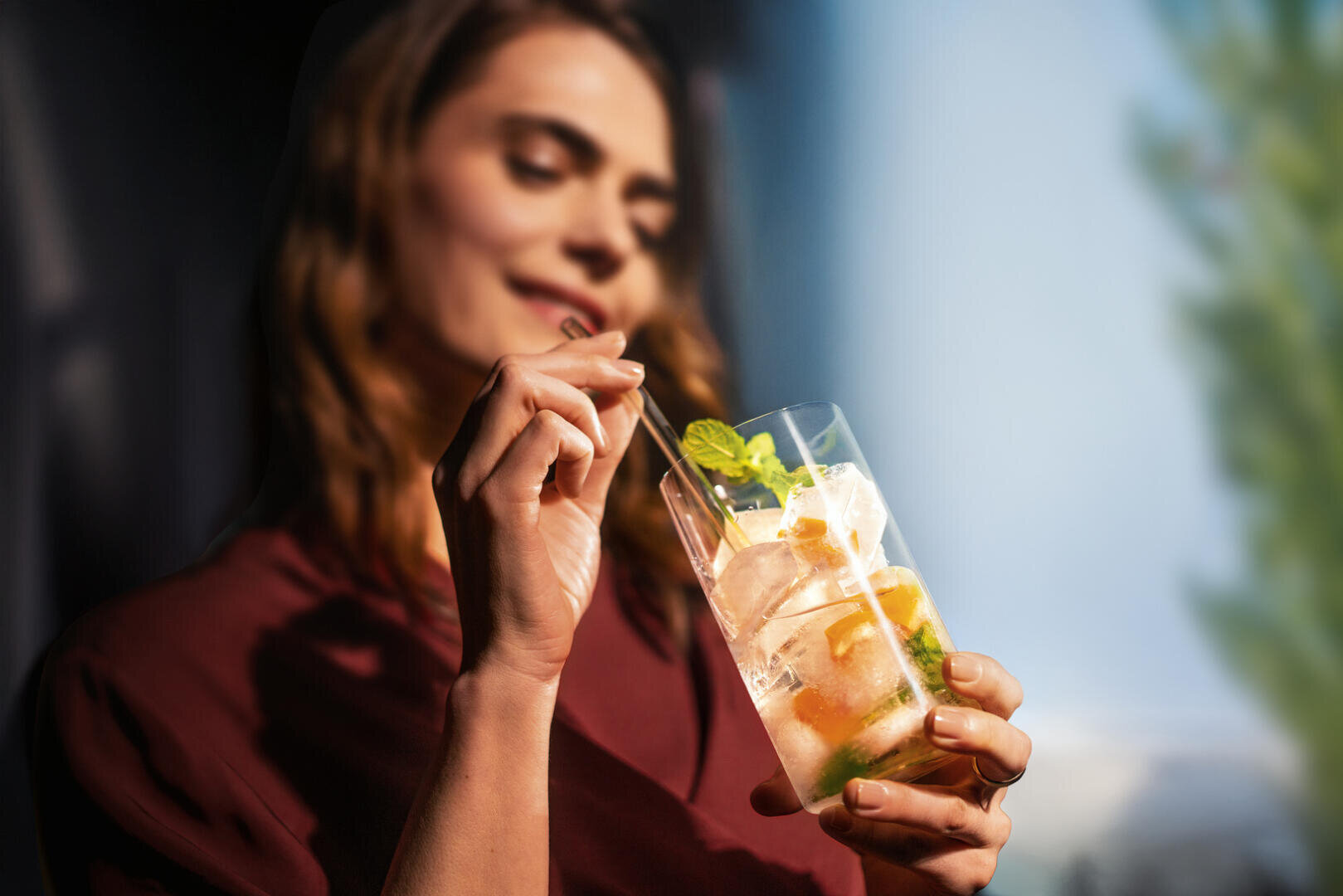 Eine Frau in kastanienbrauner Kleidung hält ein Glas mit einem eisgekühlten Getränk mit Minzblättern und Limette in der Hand und rührt es mit einem Strohhalm um, während sie das Glas ansieht und sanft lächelt.