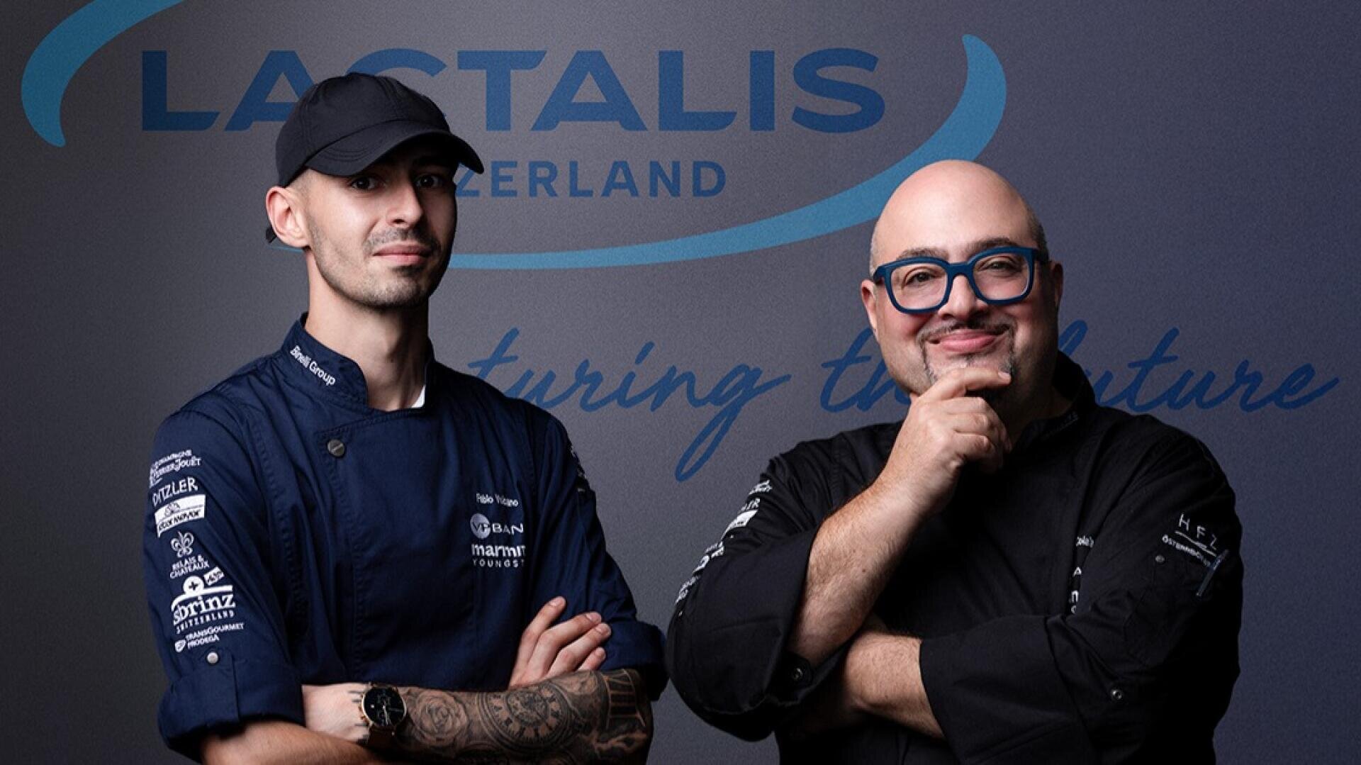 Deux chefs cuisiniers posent devant un mur portant le logo de Lactalis Suisse ; l'un porte une casquette noire et un uniforme bleu marine avec des tatouages visibles, tandis que l'autre, chauve avec des lunettes, sourit, la main sur le menton, vêtu de noir.