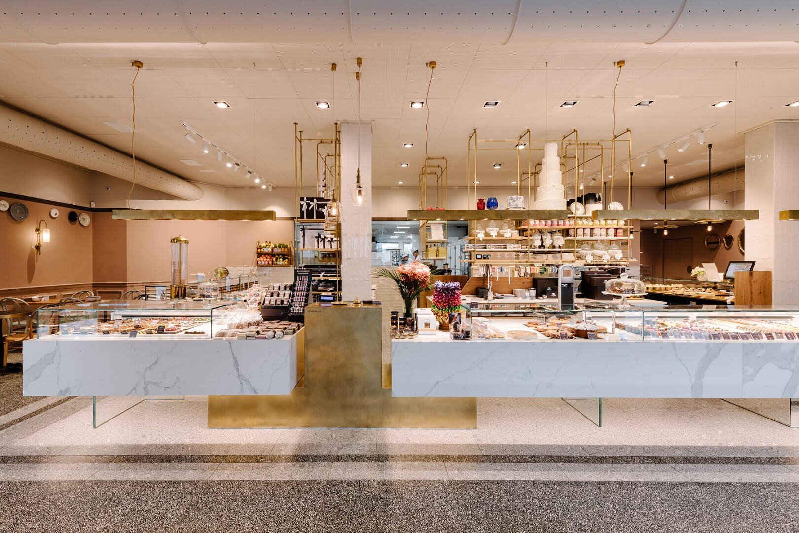 Moderne Bäckerei mit Marmor- und Goldakzenten, die eine Vielzahl von Backwaren, Kuchen und Broten in Vitrinen ausstellt. Regale mit Gläsern und Schachteln säumen die Rückseite, und frische Blumen schmücken den zentralen Tresen.