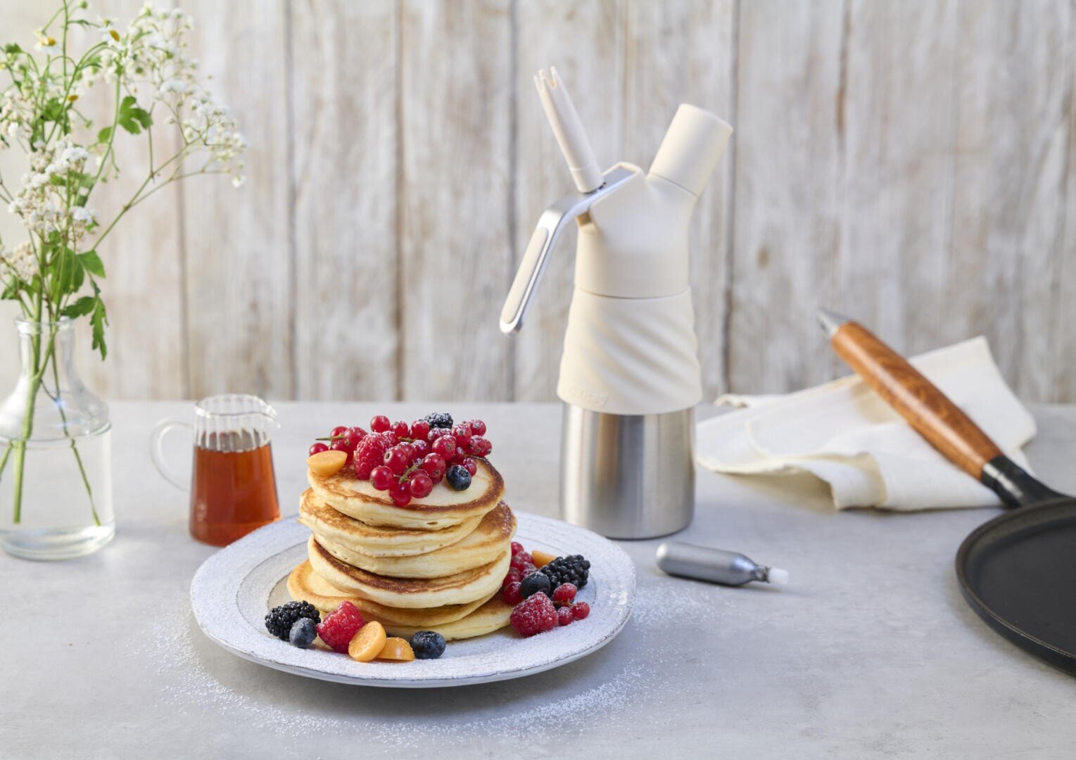 Une pile de crêpes garnies de baies fraîches est posée sur une assiette blanche, entourée de plus de baies et de sirop, à côté d'un distributeur de crème fouettée, d'une poêle, d'un torchon et d'un vase avec des fleurs sur une table lumineuse.