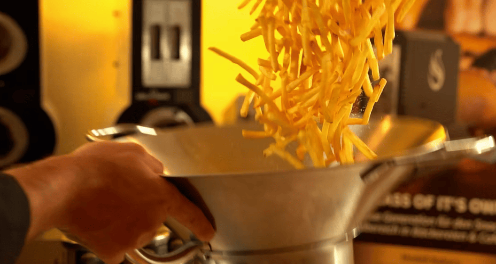 Une personne jette une charge de frites dans un grand bol en métal, les frites s'envolant vers le haut sur un fond chaud et doré.