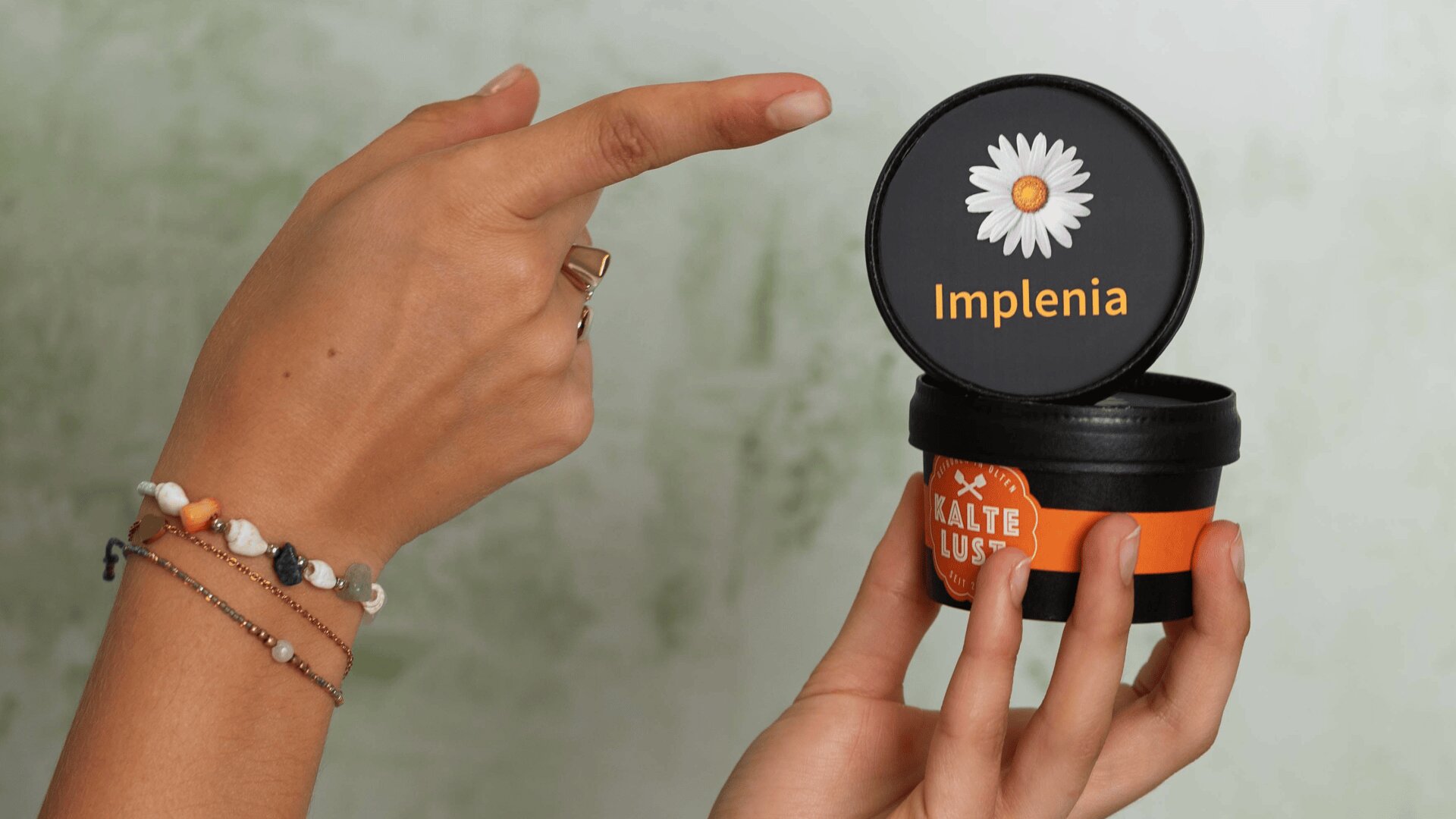 Une personne tient un petit récipient noir avec une étiquette orange et montre le couvercle, qui affiche une marguerite blanche et le mot Implenia en texte jaune. La personne porte des bracelets et une bague.