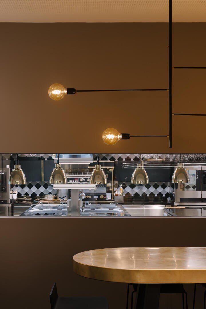 Cuisine moderne avec un mur brun, deux luminaires à ampoules apparentes et une fenêtre montrant une cuisine professionnelle en acier inoxydable avec des lampes en laiton et un dosseret à carreaux. Une table ronde se trouve au premier plan.
