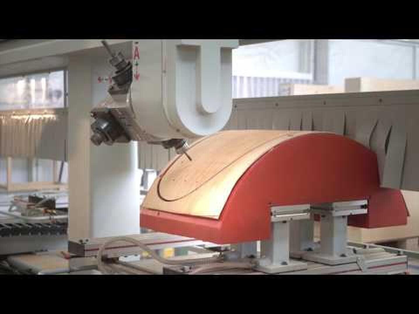 Eine CNC-Maschine schnitzt oder formt ein gebogenes Holzstück, das mit Zangen an Ort und Stelle gehalten wird. Der Arbeitsbereich ist innenliegend und gut beleuchtet.